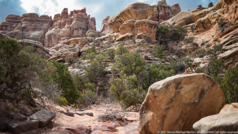 A large boulder borders a rough canyon path that descends through shrubs beneath layered cliffs.