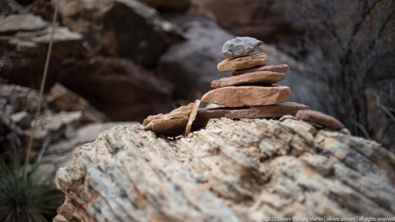 Flat tan, red, and gray stones form a small cairn on a pale outcrop, with boulders blurred behind.