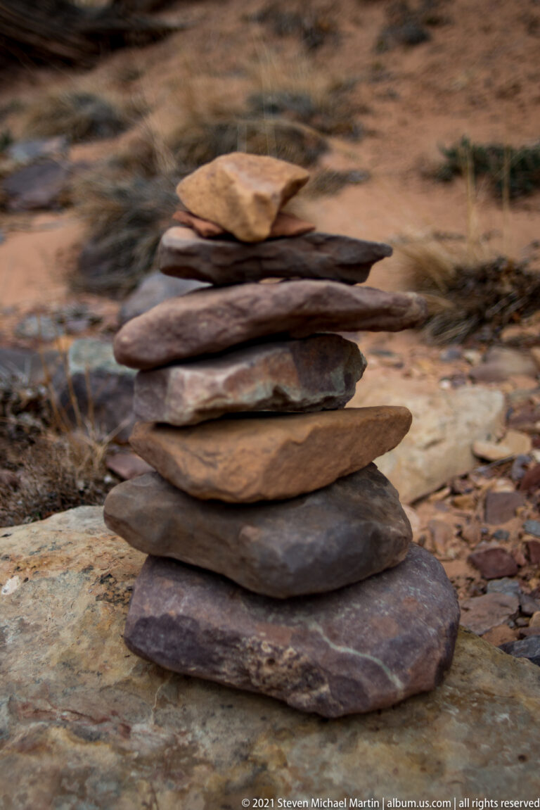 Flat red, brown, and tan stones form an uneven cairn on a boulder, topped by a small pale stone.