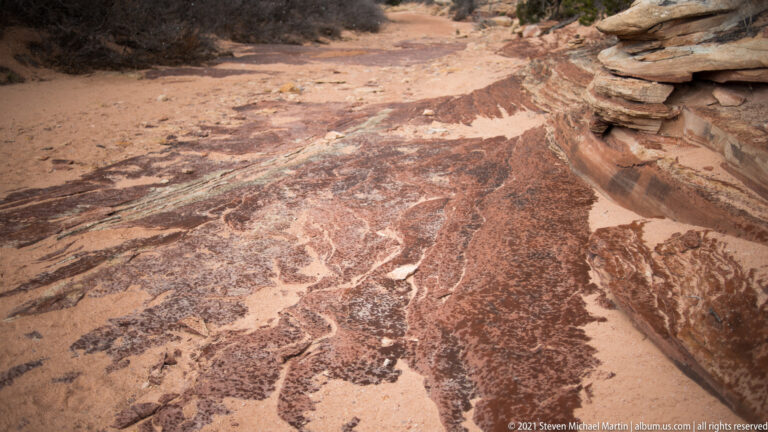 Reddish-brown staining and pale winding channels cross a sloping sandstone surface, with a small stone stack at upper right.
