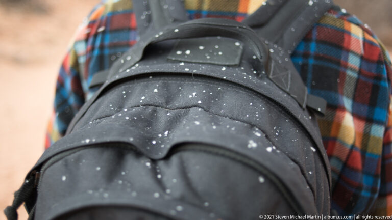 Water droplets cover a black hiking backpack worn over a colorful plaid shirt.