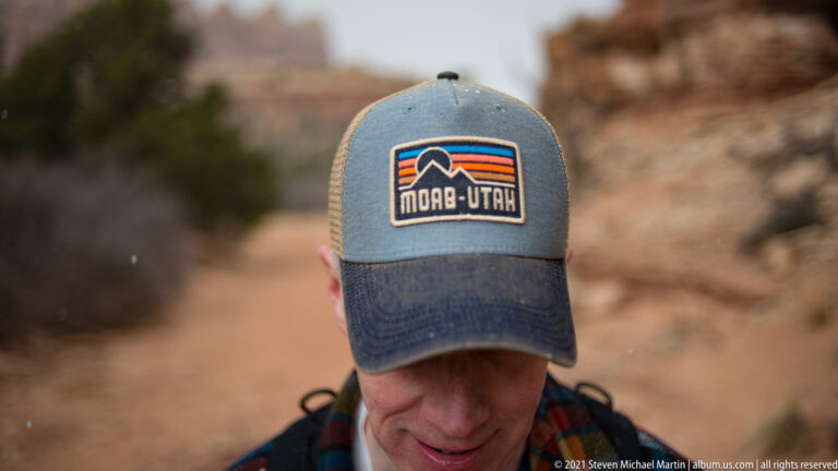 A hiker looks down beneath a blue cap bearing a Moab Utah patch, with canyon rock blurred behind.