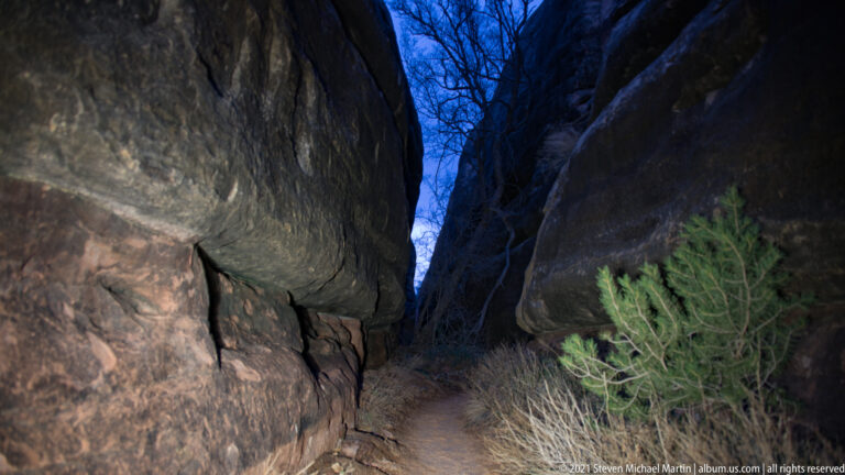 A large sandstone boulder overhangs a narrow trail between rock walls, with bare branches against blue sky above.