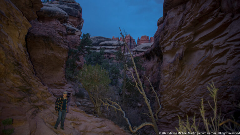 A hiker stands beside a narrow rocky trail between sandstone walls, with brush and distant formations at dusk.