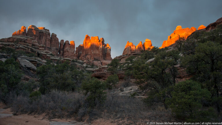 Orange-lit sandstone spires rise above a shadowed, brush-covered slope beneath a cloudy sky.