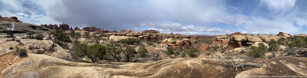 Pale slickrock slopes toward red sandstone ledges and distant pinnacles beneath a partly cloudy sky.