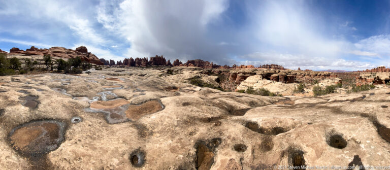 Dark shallow basins dot pale slickrock in front of red sandstone towers and a cloud-filled sky.
