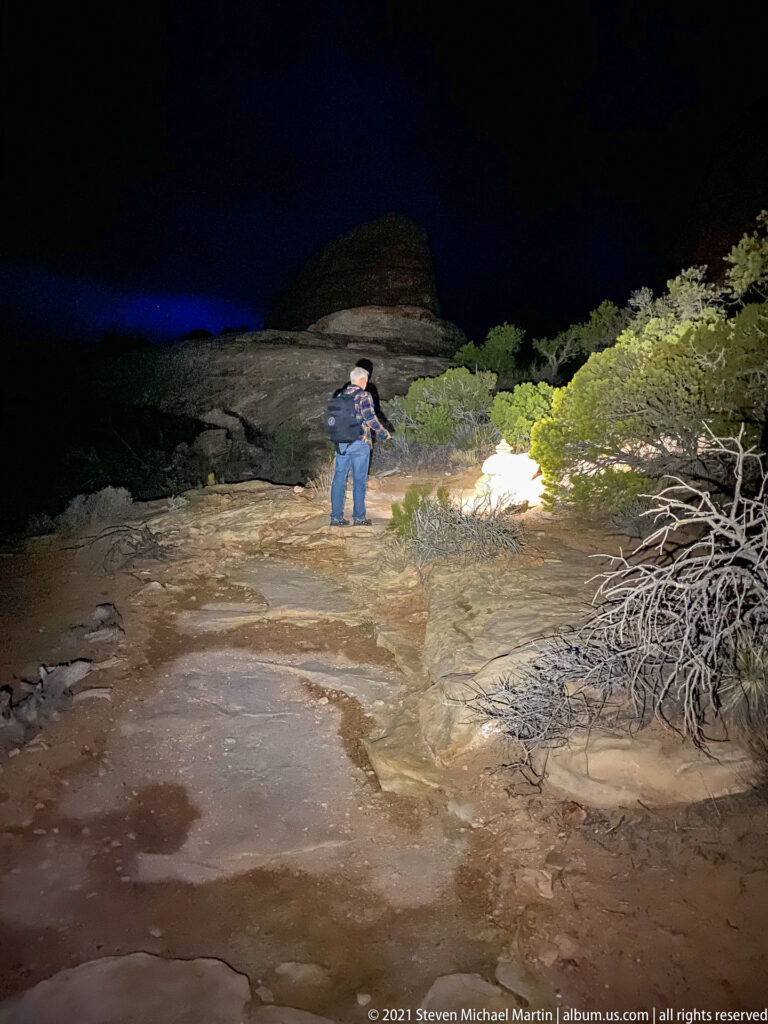 A hiker stands on a flashlight-lit sandstone shelf beside shrubs, with a low rock rise beneath a dark night sky.