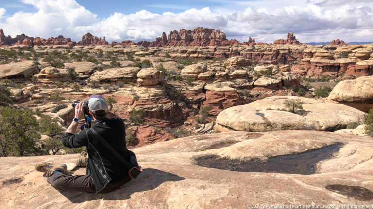 A seated photographer aims a camera toward a panorama of slickrock and distant sandstone pinnacles.