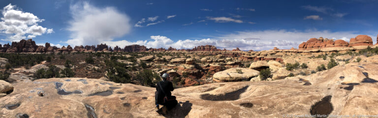 A hiker crosses pitted pale slickrock toward distant red pinnacles, casting a long shadow.