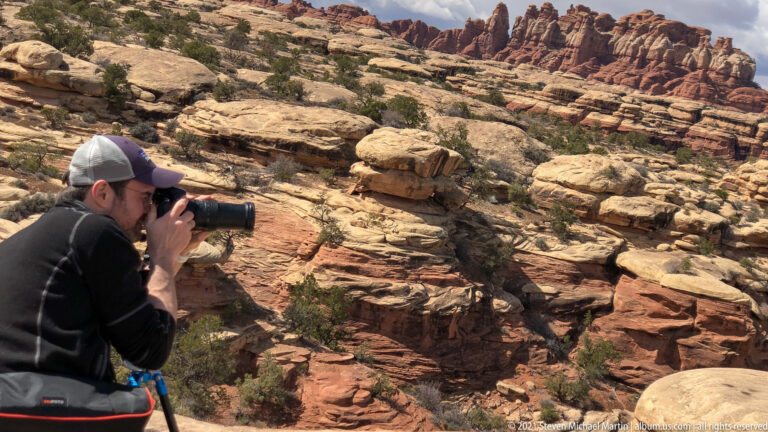 A crouching photographer aims a camera across layered sandstone ledges and a scrub-filled ravine.