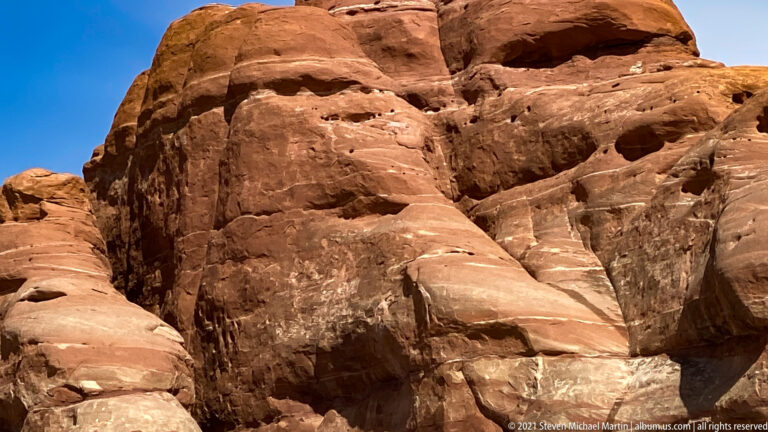 Close-up of a red sandstone wall with horizontal layers, cavities, ledges, and a dark vertical seam.
