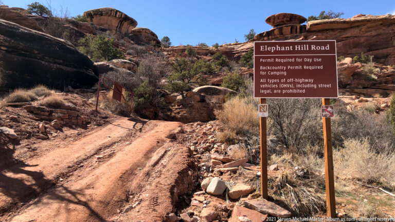 A brown Elephant Hill Road sign lists permit requirements beside a rocky dirt track in sandstone terrain.