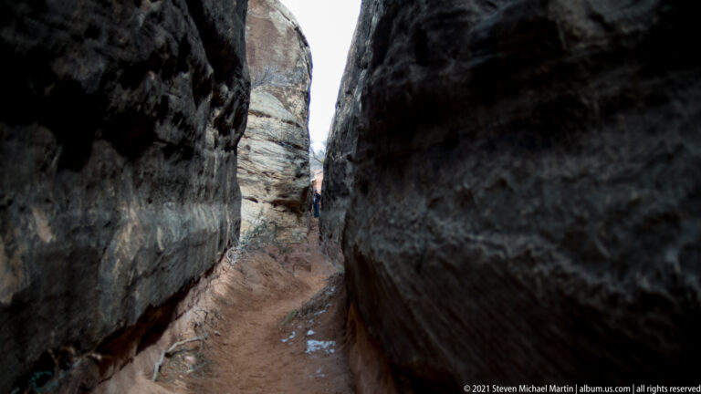 A tiny hiker stands at the far end of a winding dirt path between tall sandstone walls.