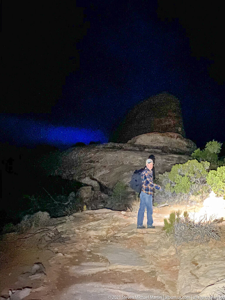 A hiker in a plaid shirt and backpack stands on slickrock at night, extending an arm toward shrubs under a dark sky.