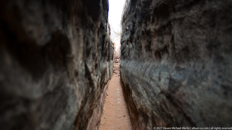 A thin reddish path recedes between tall dark sandstone walls toward a bright opening.