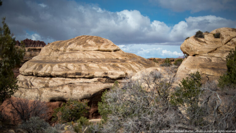 A rounded sandstone outcrop rises above scrub beneath clouds, with taller red formations in the distance.