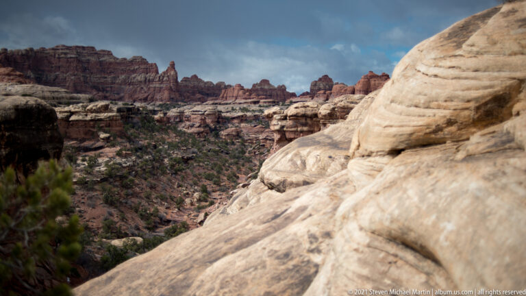A pale slickrock ledge overlooks juniper slopes, red mesas, and distant sandstone towers.