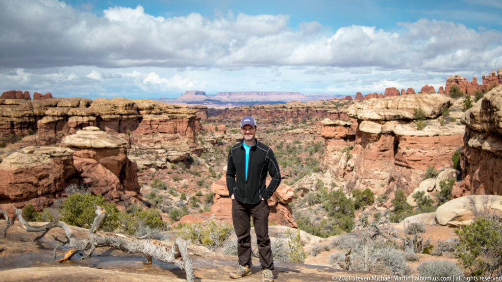 A visitor stands on a slickrock overlook above a broad canyon, with a fallen branch in the foreground.
