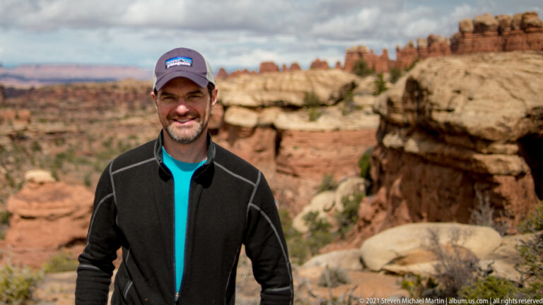 A smiling hiker in a cap and dark jacket stands before a softly blurred red-rock canyon.