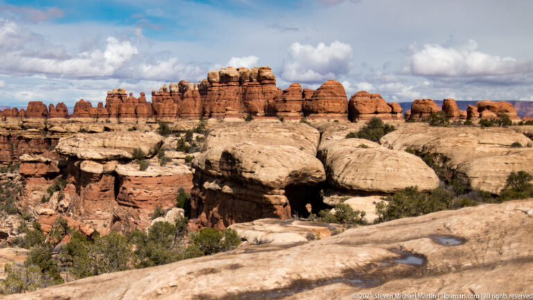 Rounded banded sandstone towers stand beyond a pale ledge with a dark opening and small pool.