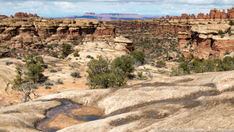 A shallow pool lies in foreground slickrock above a broad canyon of red mesas and sandstone benches.