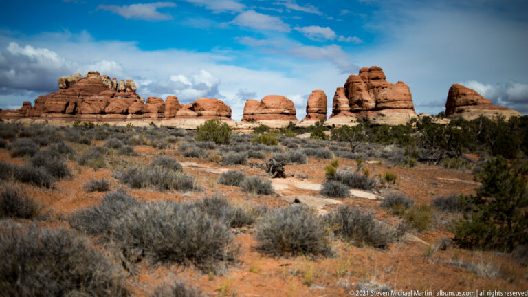 Rounded red sandstone towers line the horizon beyond a broad scrub flat under scattered clouds.