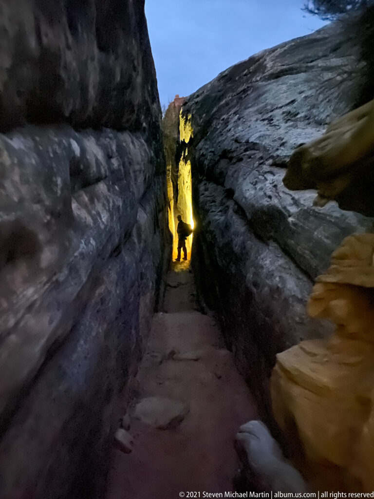 A silhouetted hiker walks through a narrow sandstone cleft toward a bright opening between high rock walls.