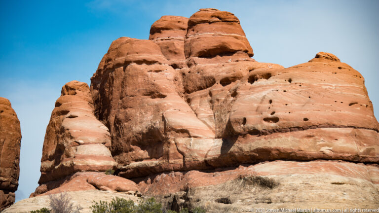 Rounded sandstone towers show dark horizontal seams and weathering pits beneath a narrow band of blue sky.