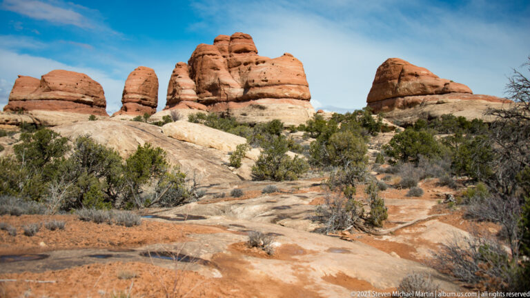 A tall rounded tower and smaller cap-shaped formation rise beyond pale shelves and juniper.