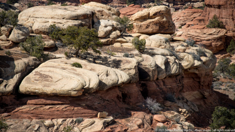 Pale rounded ledges project over a shadowed red-rock drop, with junipers growing from cracks.