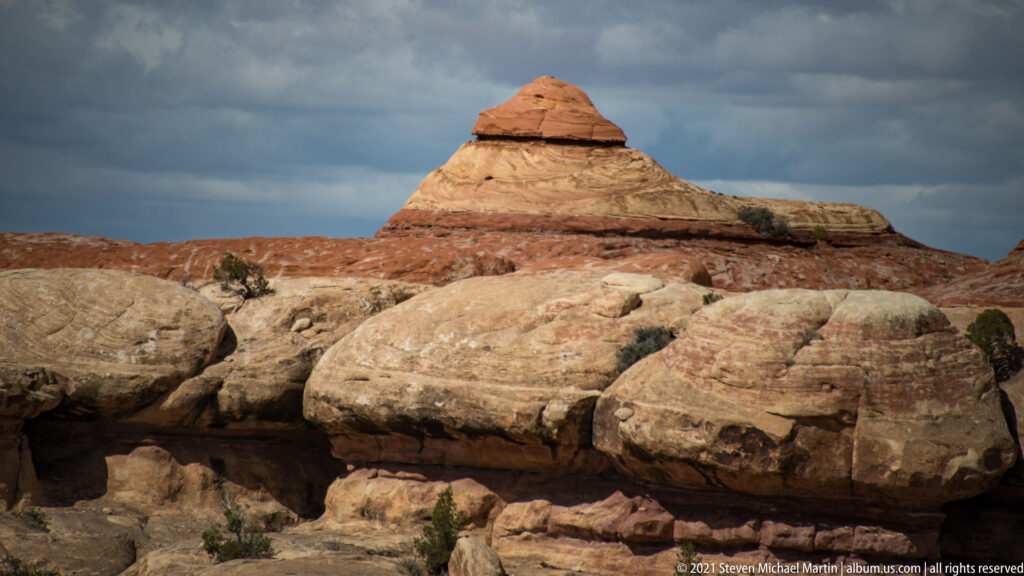 A cap-shaped orange summit rises above a pale sandstone bench beneath dark clouds.
