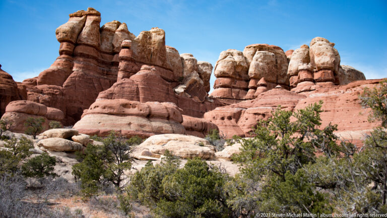 Banded red and cream towers rise above juniper and scrub beneath a clear blue sky.