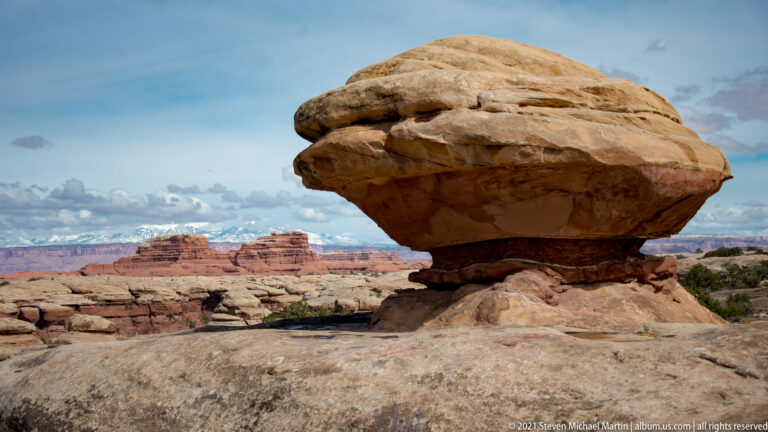 A broad rounded boulder rests on a narrow pedestal above a sandstone platform, with mesas behind.
