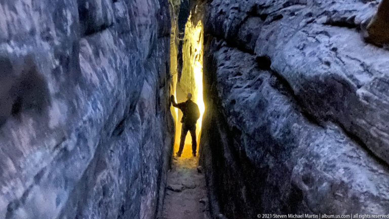 A silhouetted person touches a sandstone wall inside a narrow fissure lit by a bright vertical opening.