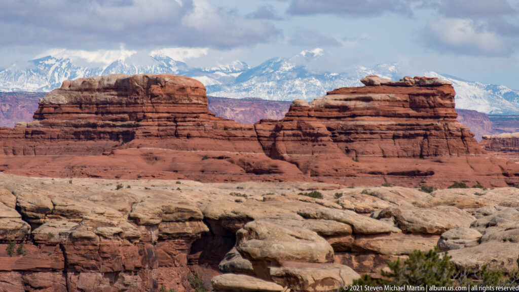 Red layered mesas stand in front of distant snow-covered peaks above pale sandstone ledges.