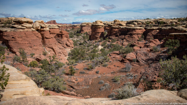 A scrub-lined ravine descends between pale sandstone shelves toward red mesas beneath broken clouds.