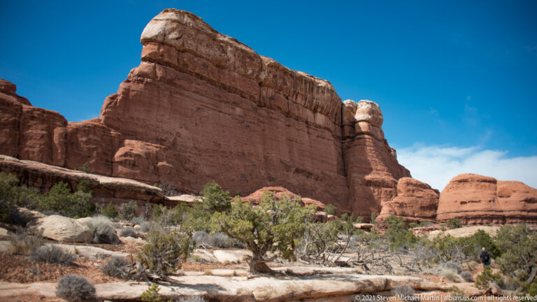 A lone hiker stands at far right below a tall, banded red cliff and scrub-covered slope.
