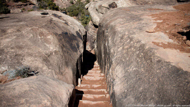 A narrow stairway descends through a dark cleft between close rounded sandstone walls.
