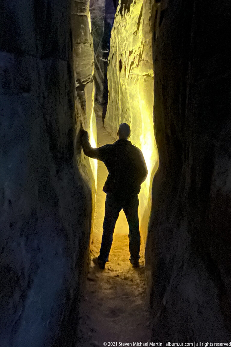 A silhouetted hiker braces between sandstone walls in a narrow passage glowing with warm light.