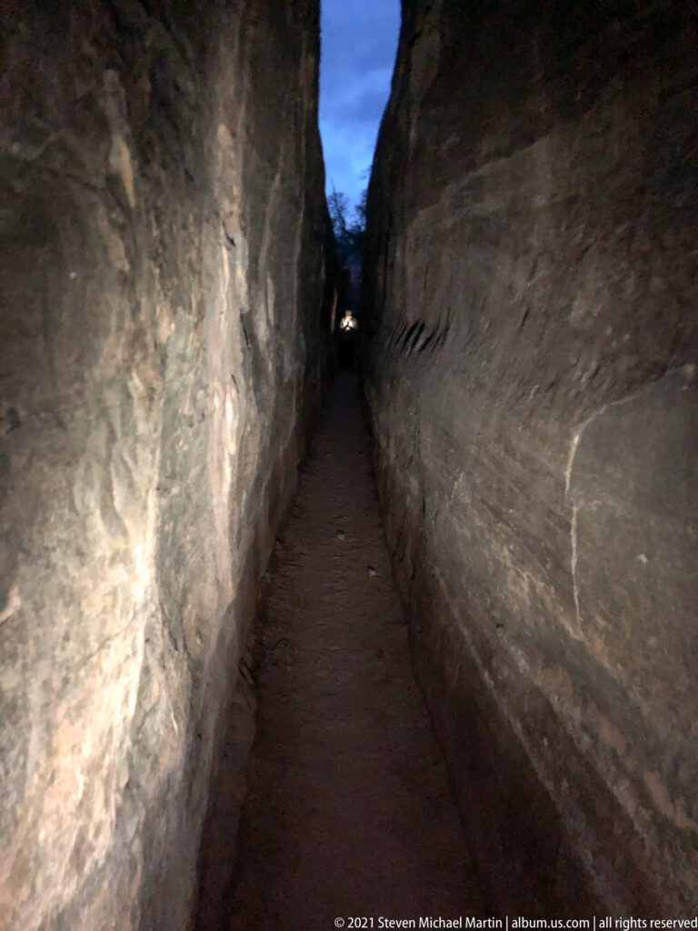 An empty dirt path recedes between tall sandstone walls toward a small opening of blue evening sky.