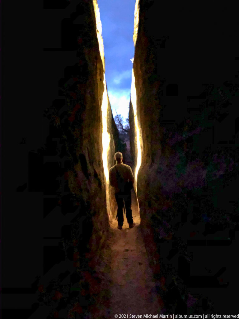 A person walks away through a narrow sandstone cleft toward a bright opening beneath blue sky.
