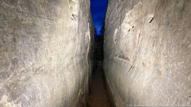 A narrow dirt path runs between high pale sandstone walls, with a thin strip of deep blue sky above.