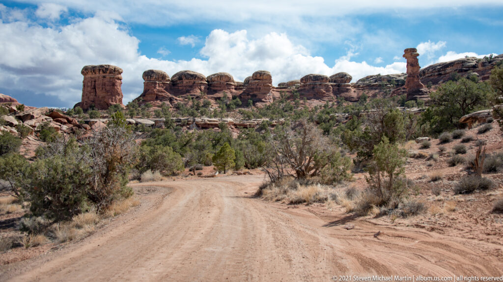 A dirt road curves through scrub toward sandstone towers beneath a cloudy blue sky.