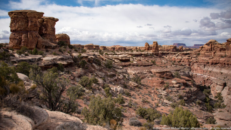 A tall sandstone tower at left overlooks a wide basin of ledges, shrubs, and distant mesas.