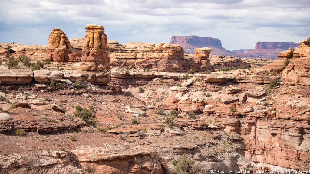 Numerous sandstone pinnacles rise beyond pale ledges and shrubs, with distant mesas beneath clouds.