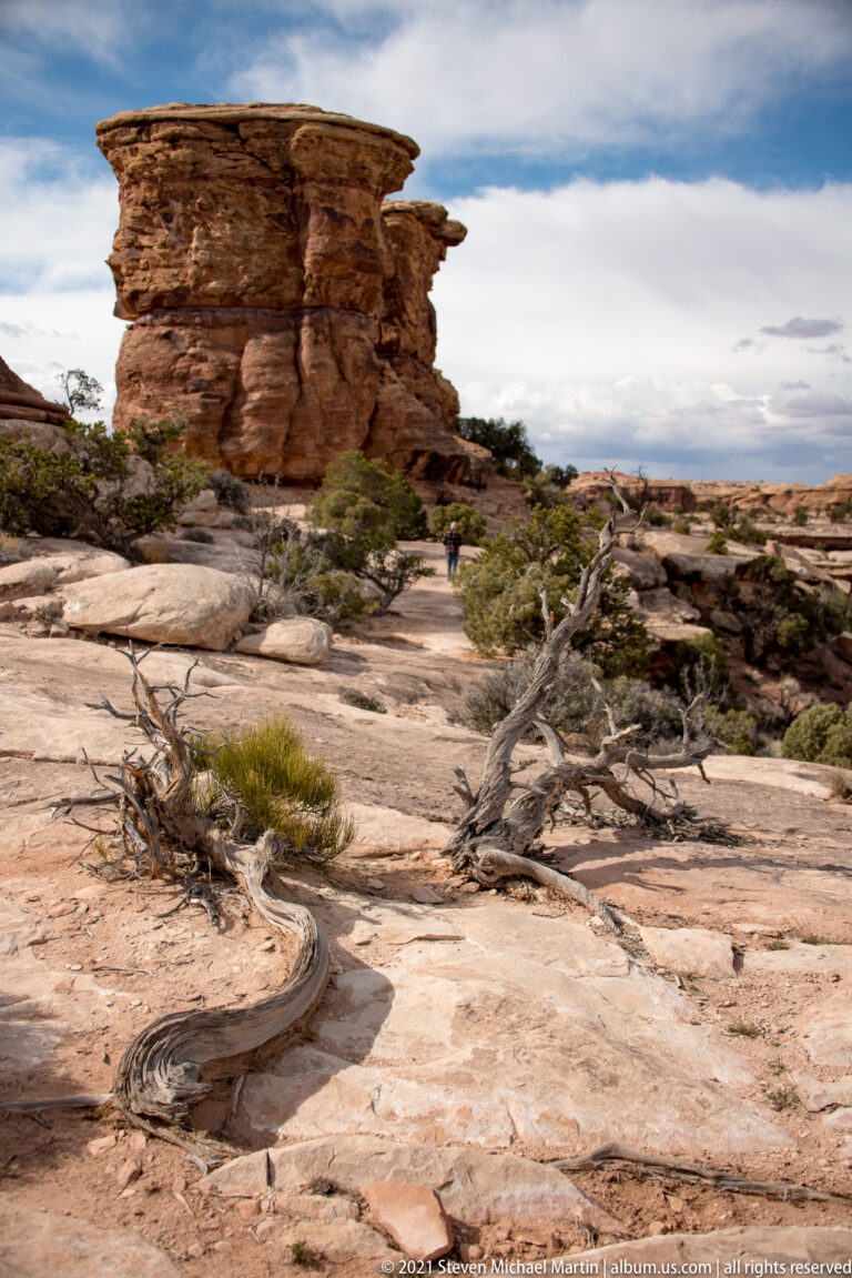 A small hiker walks across pale slickrock beneath a broad sandstone overhang and sparse shrubs.