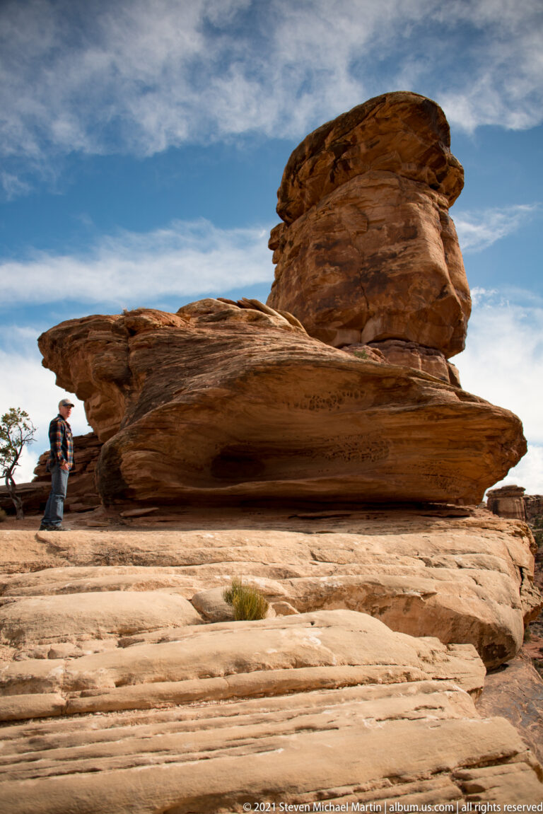 A hiker stands beside a broad mushroom-shaped sandstone formation with a projecting caprock.