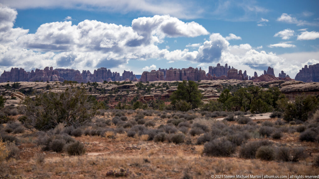 A distant line of sandstone pinnacles rises beyond a flat scrub-covered plain beneath bright clouds.