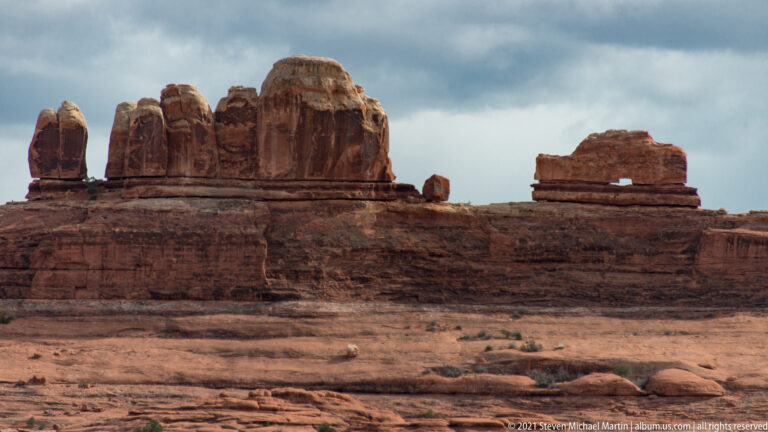 A long layered red cliff supports a row of narrow pinnacles above a pale rock shelf.